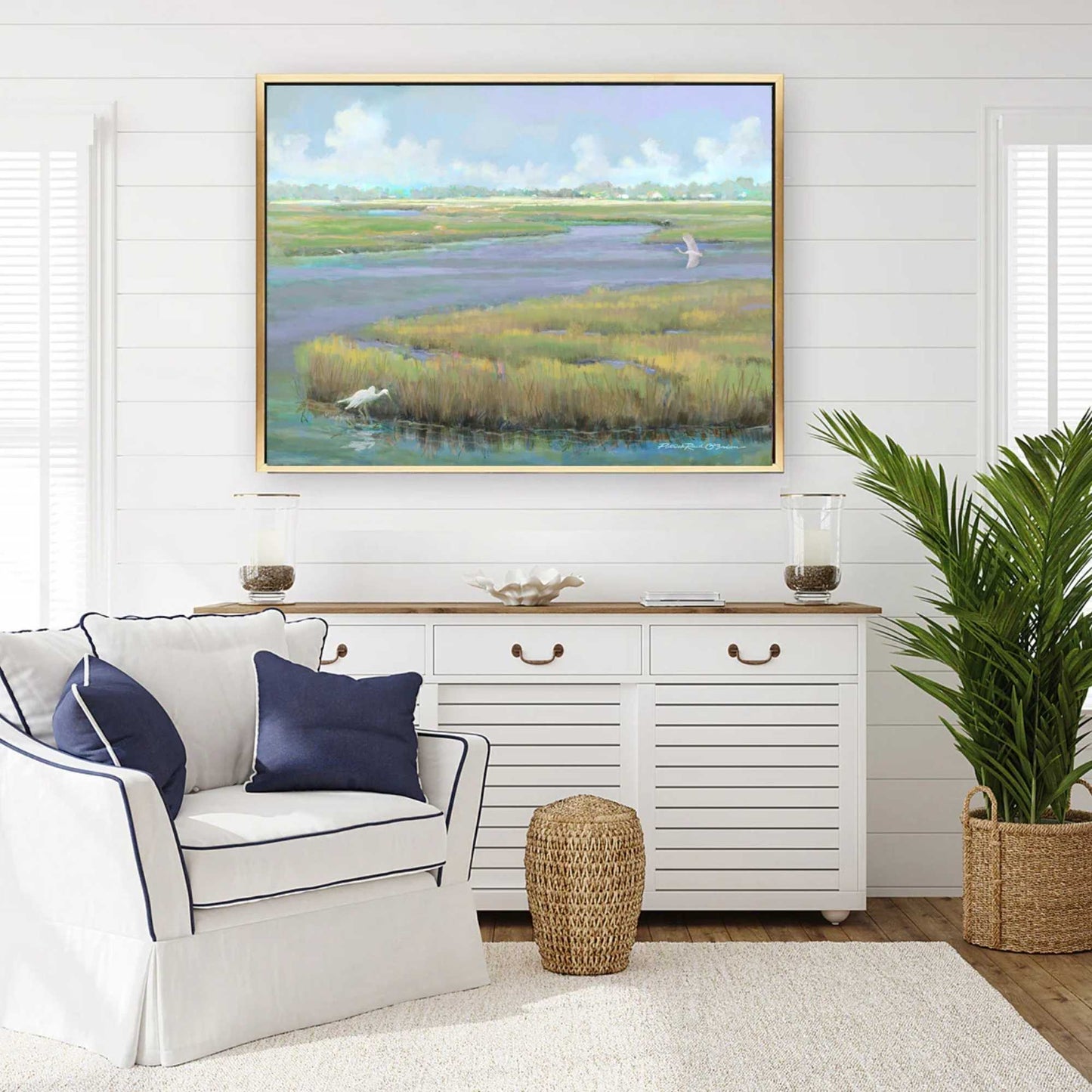 Living room with a painting of a coastal marsh landscape on the wall, white sofa with blue pillows, and a sideboard.